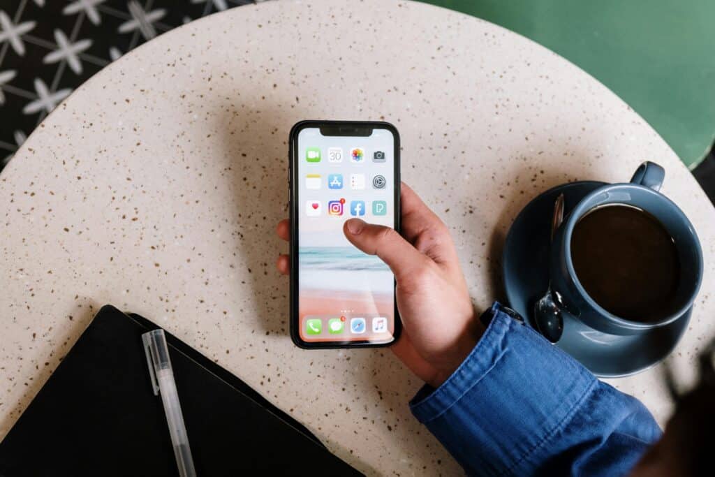 A hand holding a phone with social media icons on the screen next to a cup of coffee on a tan table.